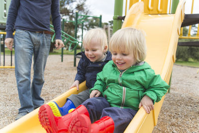 Low section of father standing by sons playing on slide at playground