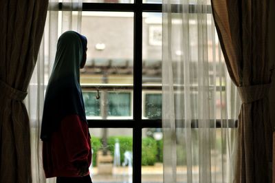 Side view of boy wearing hooded shirt looking through window at home