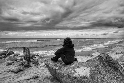 Full length of person sitting on rock at beach against cloudy sky