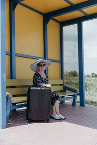 An elderly woman with a suitcase sits at a bus stop.