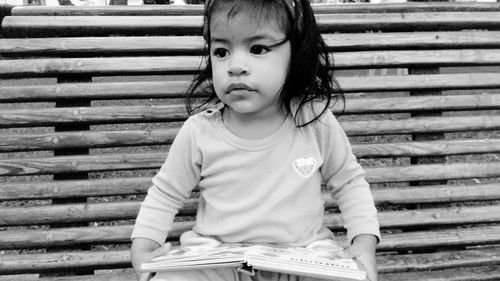 Cute baby girl holding book looking away while sitting on bench