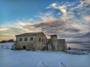 Buildings on snow covered field against sky during sunset