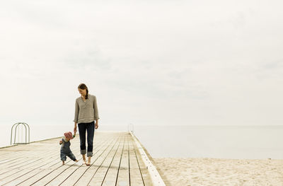 Mother and daughter walking on pier at beach against sky