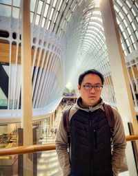 Portrait of young man standing against railing under skylight in mall.