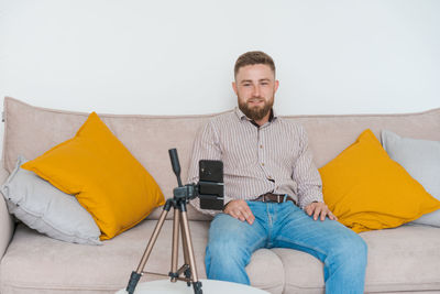 Smiling bearded guy filming video using smartphone on tripod online while