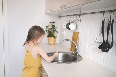 European child of four years old in yellow jumpsuit washes his hands in real bright kitchen