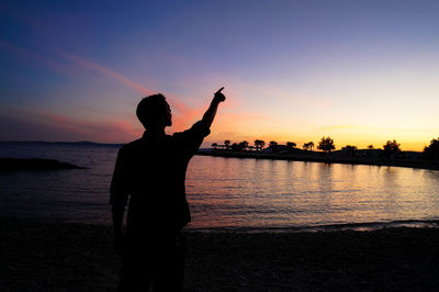 Silhouette man pointing while standing at beach against sky during sunset