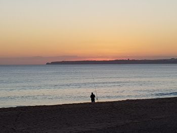 Silhouette man fishing at beach against sky during sunset