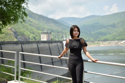 Portrait of young woman standing by railing against river