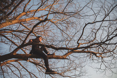 Low angle view of silhouette thoughtful boy sitting on branch against sky during sunset
