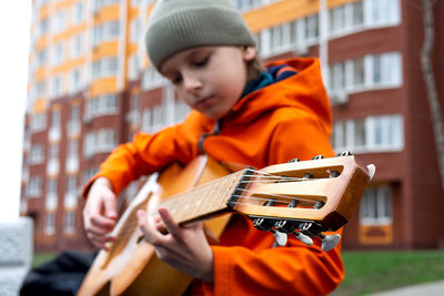 Portrait of caucasian blond boy in orange jacket playing acoustic guitar outdoors on autumn day