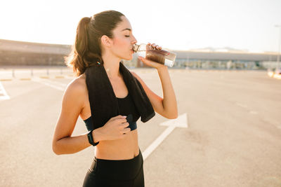 Positive young fit female in sportswear standing on parking lot and drinking refreshing beverage from bottle while resting after training