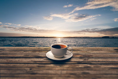 Coffee cup on table by sea against sky during sunset