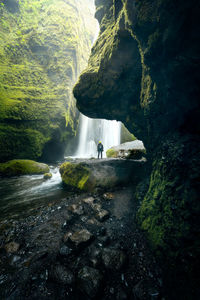 Person in moss-covered cave in front of wild waterfall and streaming river, seljalandsfoss iceland