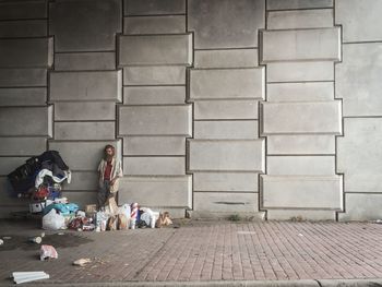 Homeless man standing by shopping cart and bags against wall