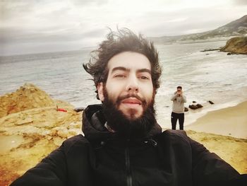 Portrait of young man standing at beach against cloudy sky