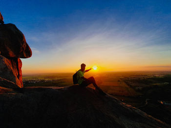 Man sitting on rock against sky during sunset