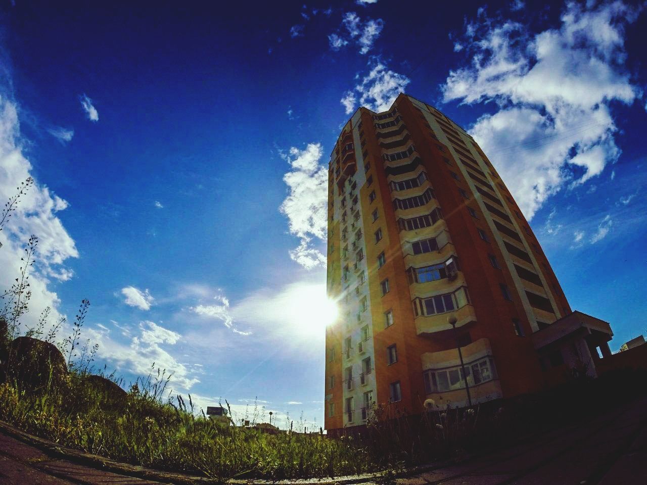 building exterior, architecture, built structure, sky, low angle view, blue, city, sunlight, cloud - sky, sun, cloud, sunbeam, building, tall - high, tree, modern, glass - material, reflection, skyscraper, lens flare