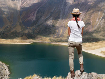 Rear view of woman standing on cliff against lake by mountains