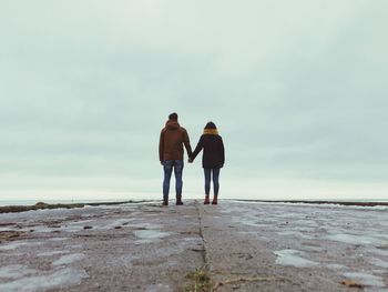 Rear view of couple holding hands while standing on pier against cloudy sky