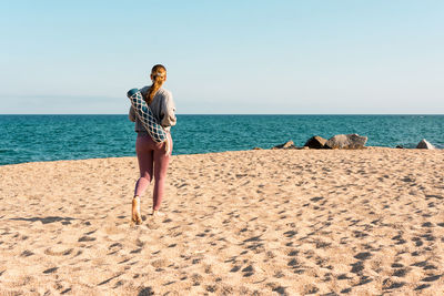 Full body back view of unrecognizable of young barefoot female in activewear with rolled yoga mat looking at distance while standing on sandy beach near sea