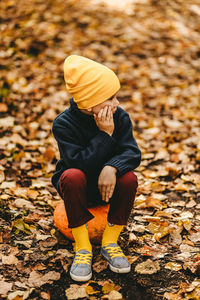 A boy a teen child in a bright yellow hat sits on a pumpkin in an autumn forest in nature outdoors