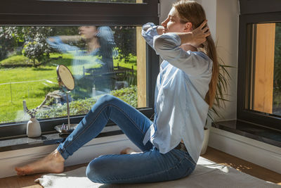 A beautiful young girl sits at home next to the window and puts on makeup and straightens her hair. 