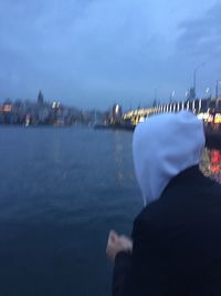 Rear view of man looking at illuminated city by sea against sky