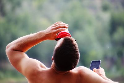 Rear view of shirtless man drinking while using mobile phone