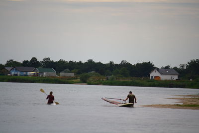 Rear view of people with paddleboard and windsurfing board in river against sky