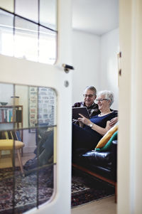 Full length of smiling senior couple sitting on sofa with digital tablet seen from ajar door