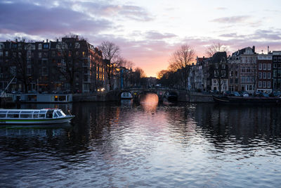 Boats moored in river by buildings against sky during sunset