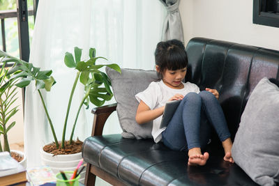 Full length of cute girl using digital tablet while sitting on sofa at home