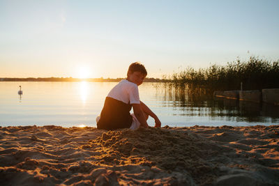 Boy sitting on beach against sky during sunset