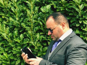 Businessman in sunglasses using mobile phone by plants