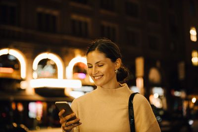 Smiling woman texting through cellphone while standing in city at night