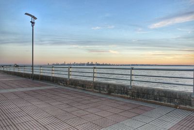 Street lights on footpath by sea against sky during sunset