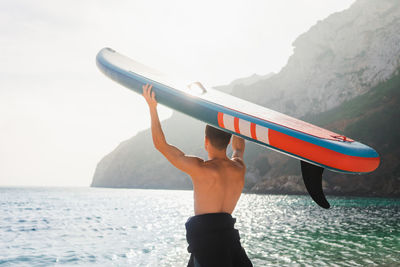Back view of unrecognizable male surfer with sup board for paddle surfing standing on pebble coast of turquoise seawater in morning