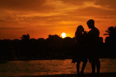 Silhouette couple standing on lakeshore against cloudy sky during sunset