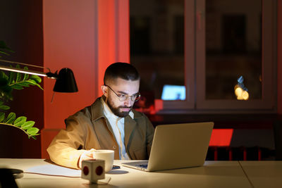 Concentrated man look at laptop drink coffee at workplace in office at dark late night overworking