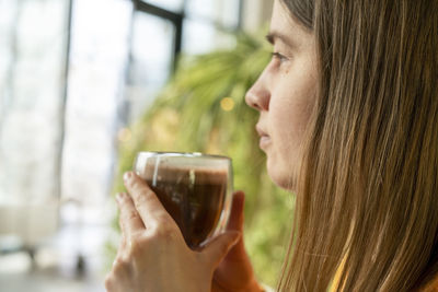 Young caucasian woman holding glass cup of coffee or cocoa and meditating looking out the window 
