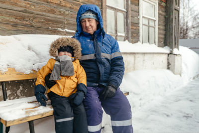 Grandpa and a little boy sit by the house on a winter day.