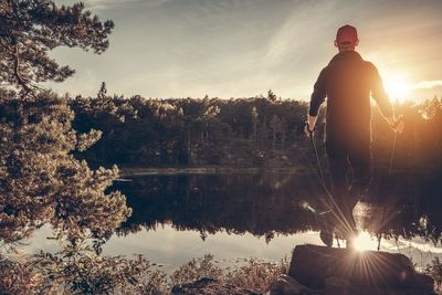 Rear view of man standing by lake against sky during sunset