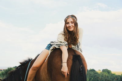 Portrait of beautiful smiling young woman sitting on brown horse on field