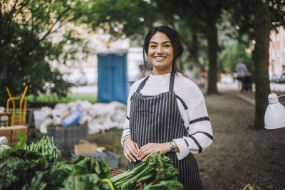 Portrait of smiling female vendor wearing apron while standing near stall at vegetable market