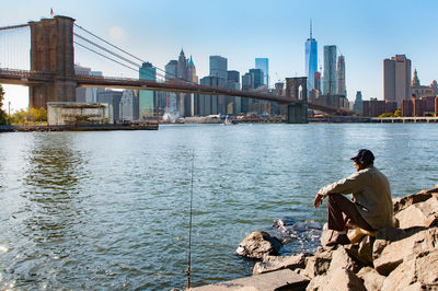 Man sitting brooklyn bridge against sky in city on sunny day