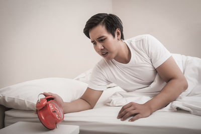 Worried young man looking at alarm clock while lying on bed