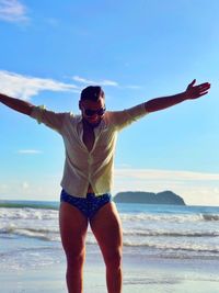 Happy man with arms outstretched standing at beach against sky on sunny day