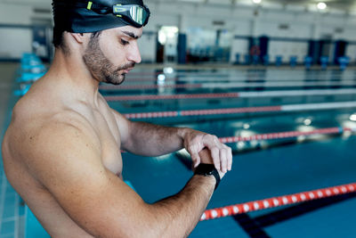 Side view of bearded male athlete in swimming cap and goggles watching time on wristwatch while on poolside