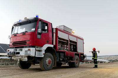 Full length of brave anonymous firefighter wearing special protective uniform and holding axe on shoulder while standing near red fire truck and looking away in countryside